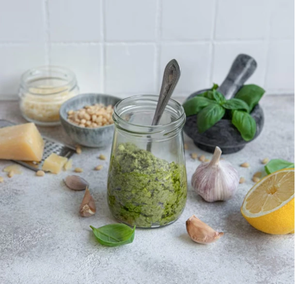 Nut-free basil pesto served in a glass jar with ingredients like basil leaves, garlic, and olive oil on a kitchen counter