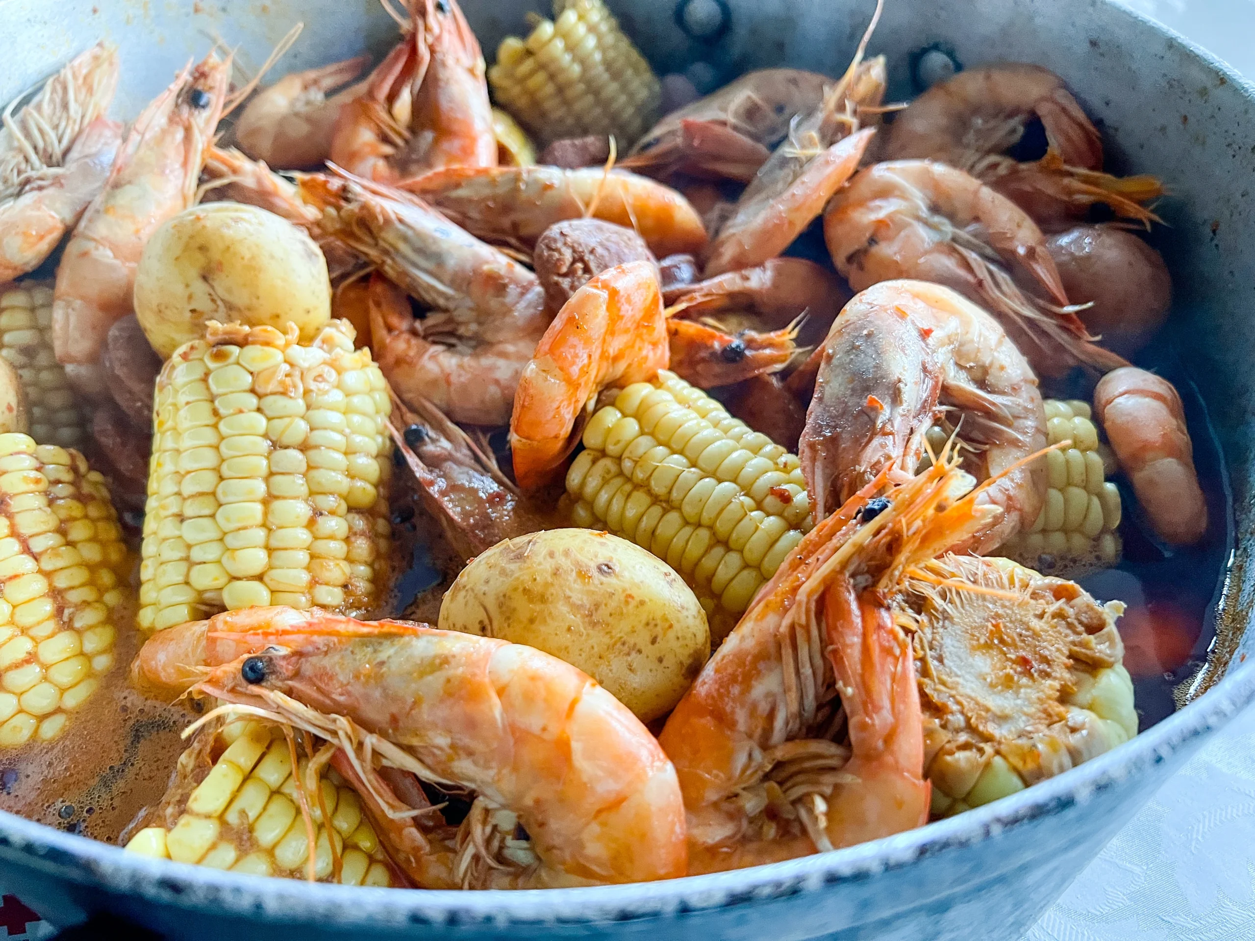 A close-up shot of a shrimp boil, featuring shrimp, corn, and potatoes, neatly arranged on a tray inside a red-and-white Igloo cooler. The cooler is lined with a checkered red-and-white cloth.