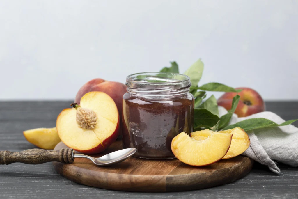 Homemade peach jam in a glass jar with fresh peaches on a wooden table.