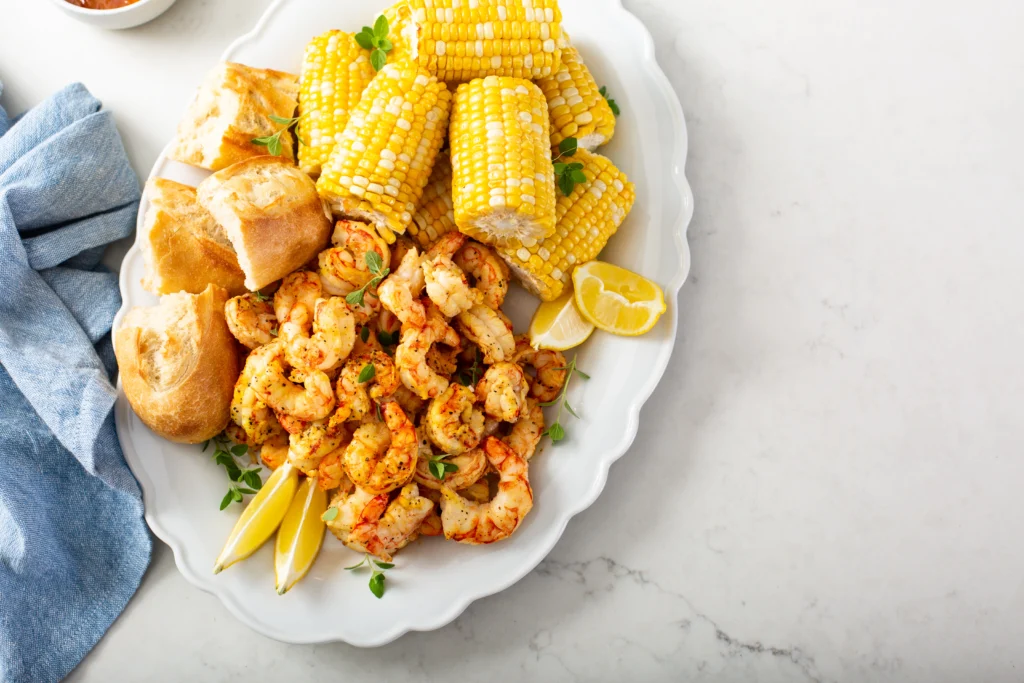 A close-up shot of a shrimp boil, featuring shrimp, corn, and potatoes, neatly arranged on a tray inside a red-and-white Igloo cooler. The cooler is lined with a checkered red-and-white cloth.