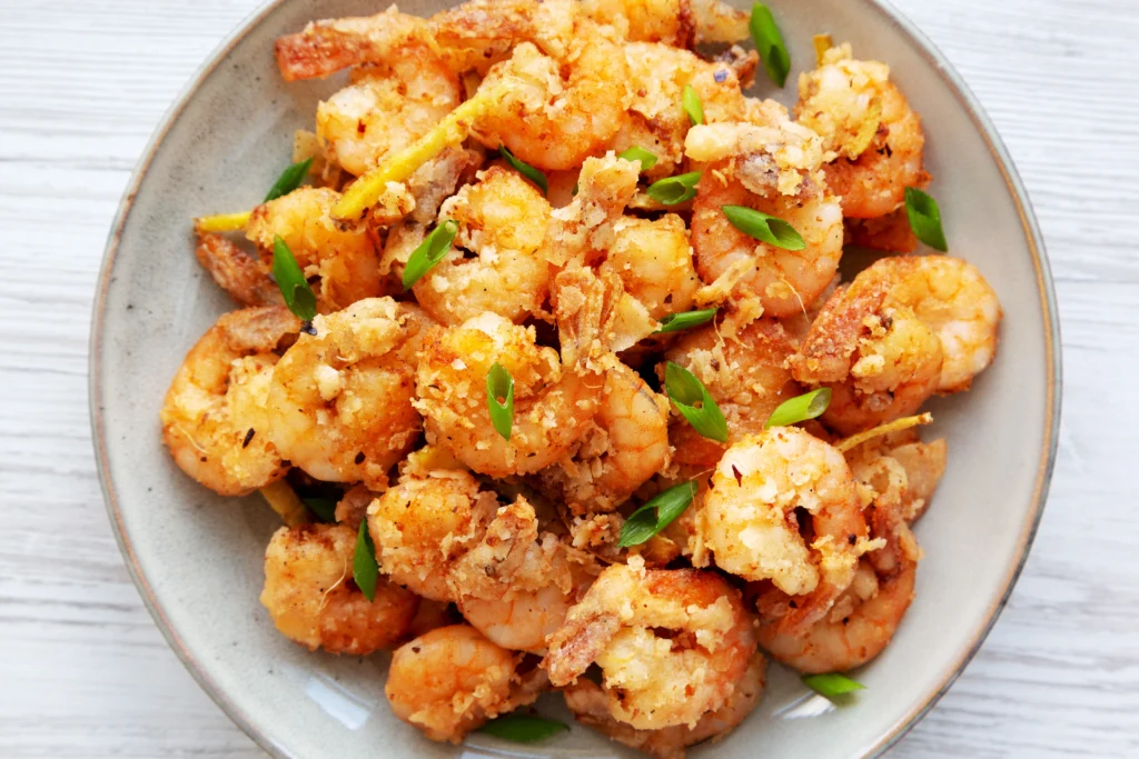 Crispy, golden-brown shrimp coated in a creamy orange sauce, garnished with finely chopped green onions, in a white bowl with an air fryer visible in the background
