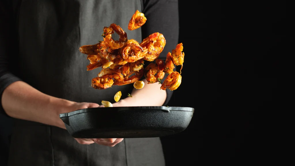 Crispy, golden-brown shrimp coated in a creamy orange sauce, garnished with finely chopped green onions, in a white bowl with an air fryer visible in the background.