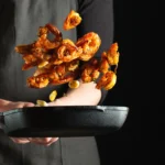 Crispy, golden-brown shrimp coated in a creamy orange sauce, garnished with finely chopped green onions, in a white bowl with an air fryer visible in the background.