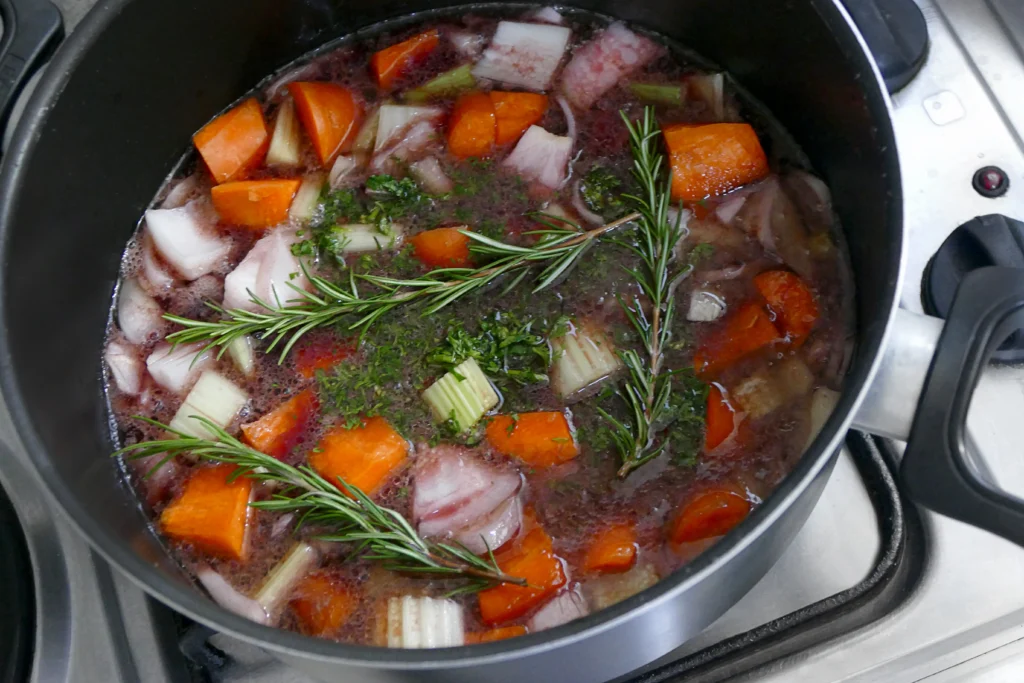 A glass jar filled with golden bone broth next to a slow cooker, surrounded by fresh herbs and vegetables.