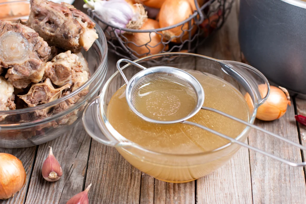 A glass jar filled with golden bone broth next to a slow cooker, surrounded by fresh herbs and vegetables.