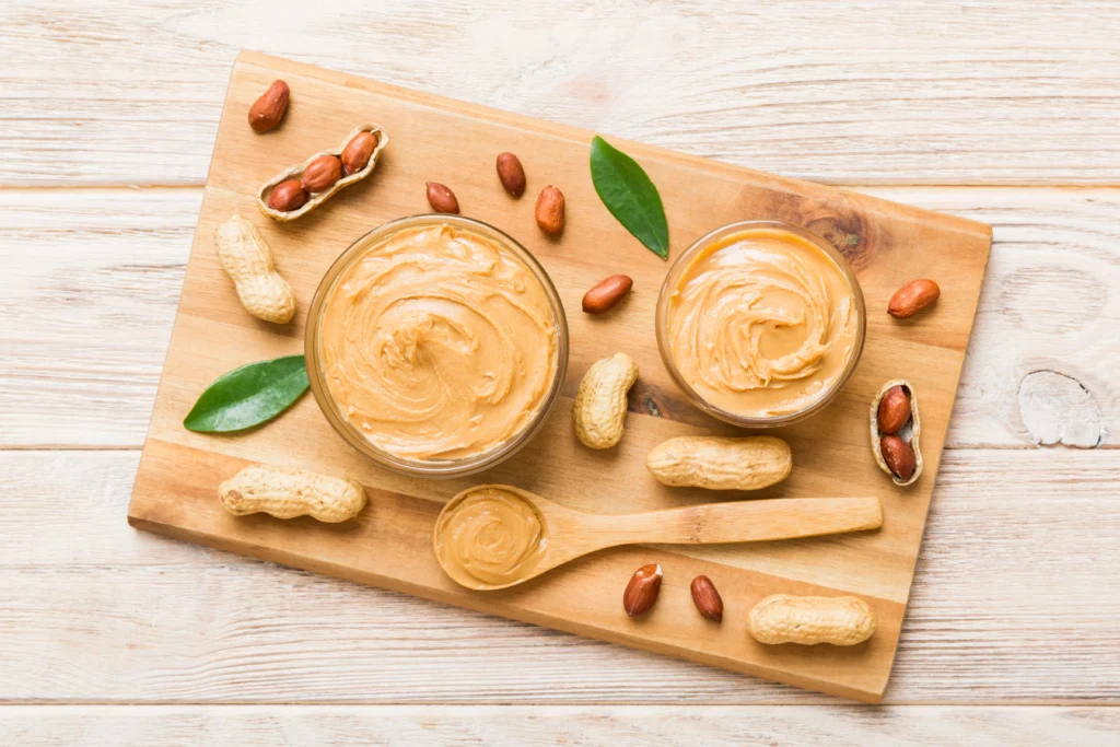 Bowl of creamy homemade peanut sauce with a spoon, surrounded by peanuts, lime, and fresh herbs.