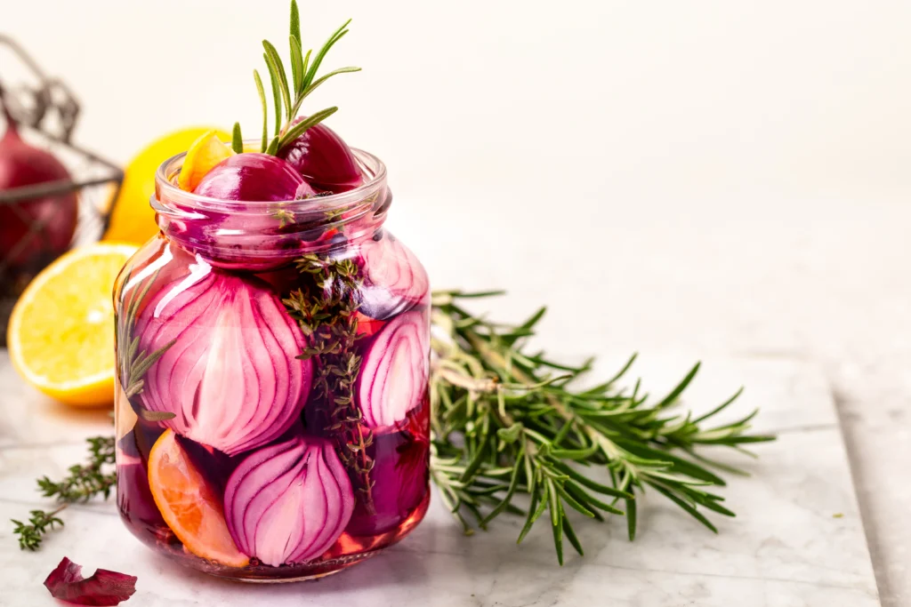 Homemade pickled red onions in a glass jar with vinegar, peppercorns, and garlic on a kitchen counter.