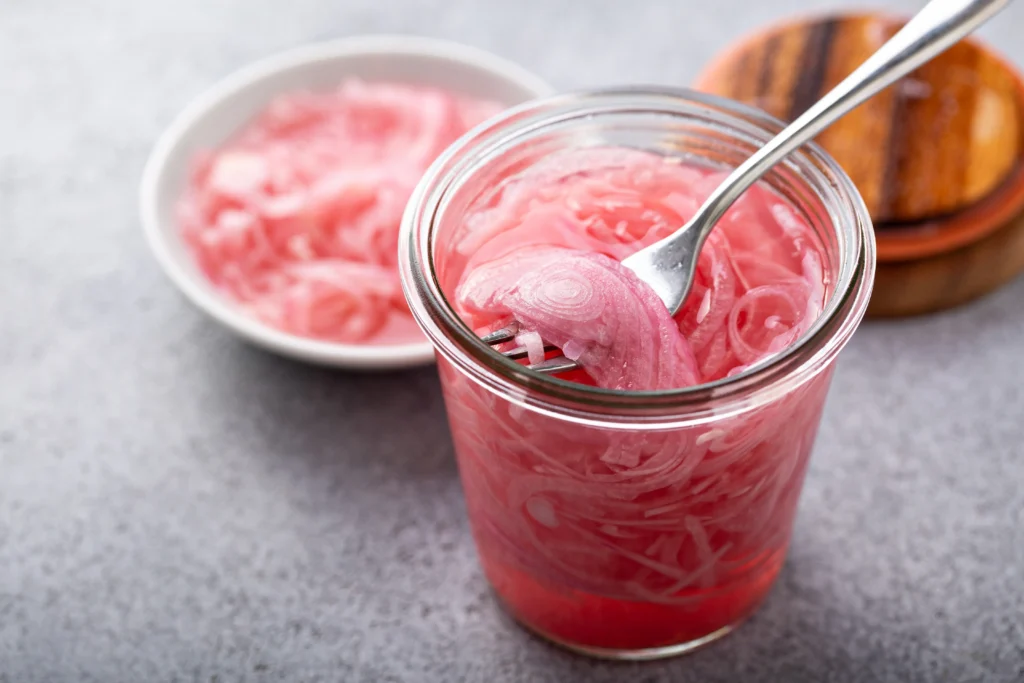 Homemade pickled red onions in a glass jar with vinegar, peppercorns, and garlic on a kitchen counter.