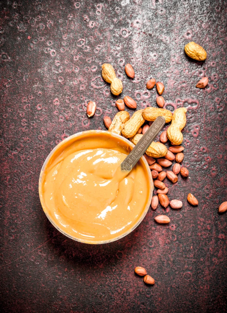 Bowl of creamy homemade peanut sauce with a spoon, surrounded by peanuts, lime, and fresh herbs.