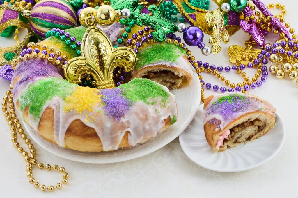 Homemade King Cake decorated with purple, green, and gold sugar icing, sliced to reveal soft cinnamon filling inside on a festive table