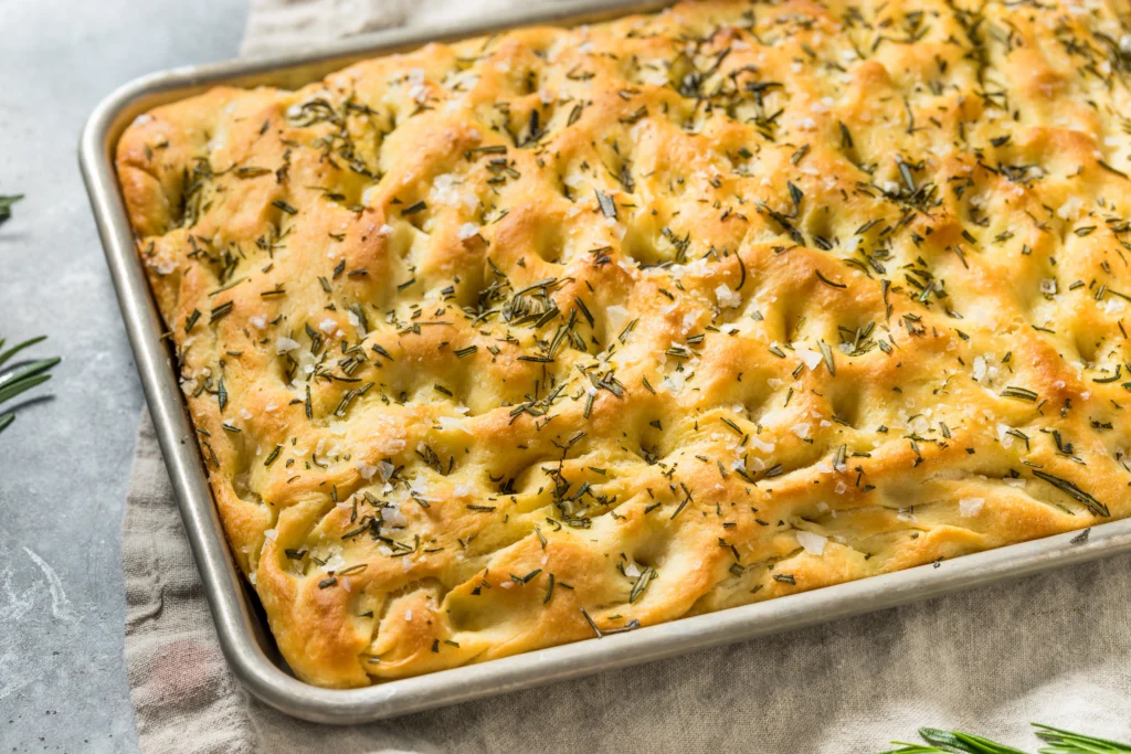 Freshly baked fluffy sourdough focaccia with airy texture, golden crispy crust, olive oil drizzle, and rosemary topping on a rustic wooden table.