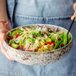 Bowl of viral crispy rice salad topped with fresh vegetables, herbs, and a creamy spicy tahini dressing, served on a wooden table.