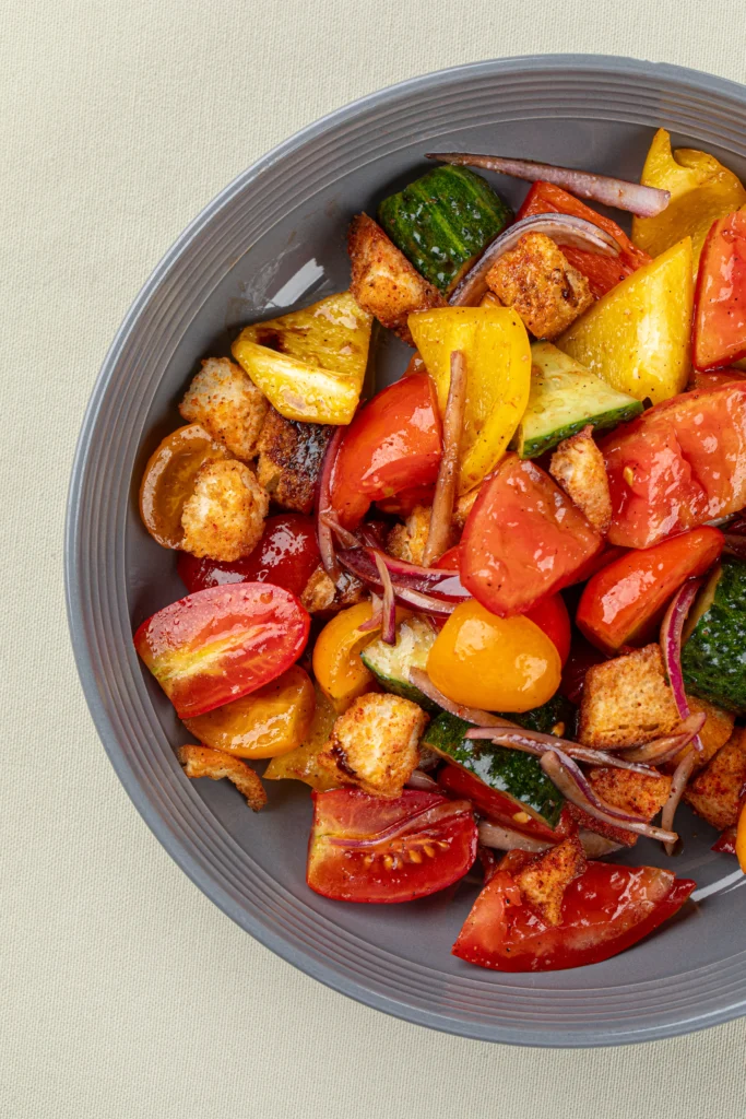 A colorful bowl of Chili Crisp Panzanella Salad made with toasted sourdough bread, ripe tomatoes, cucumbers, bell peppers, fresh basil, and a spicy chili crisp vinaigrette, garnished with herbs and served on a rustic wooden table.