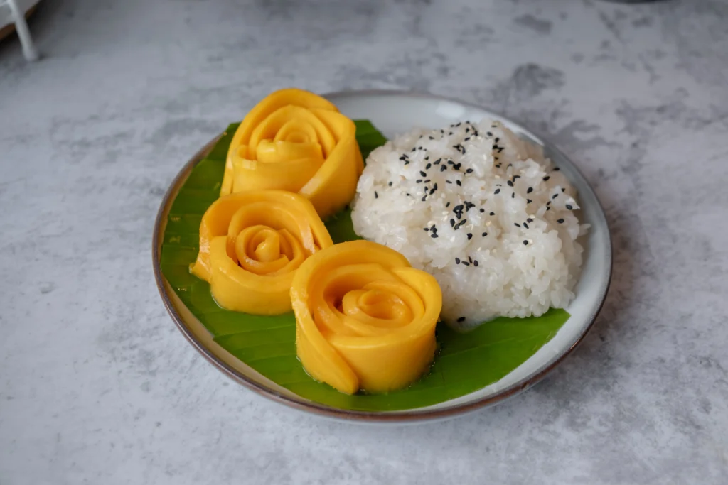 A traditional serving of sweet mango sticky rice topped with creamy coconut sauce, featuring ripe golden mango slices arranged beside glutinous rice and garnished with toasted sesame seeds on a rustic plate.