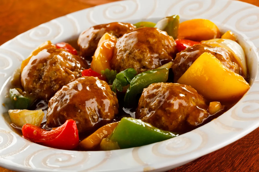 A bowl of sweet and sour meatballs coated in a glossy tangy sauce, garnished with fresh parsley and served alongside colorful bell peppers and pineapple chunks.