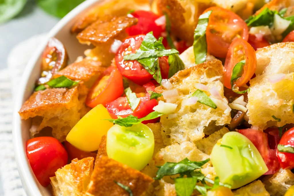 A colorful bowl of Chili Crisp Panzanella Salad made with toasted sourdough bread, ripe tomatoes, cucumbers, bell peppers, fresh basil, and a spicy chili crisp vinaigrette, garnished with herbs and served on a rustic wooden table.