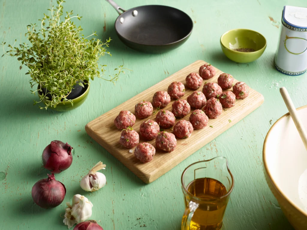 A bowl of sweet and sour meatballs coated in a glossy tangy sauce, garnished with fresh parsley and served alongside colorful bell peppers and pineapple chunks.