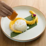 A traditional serving of sweet mango sticky rice topped with creamy coconut sauce, featuring ripe golden mango slices arranged beside glutinous rice and garnished with toasted sesame seeds on a rustic plate.
