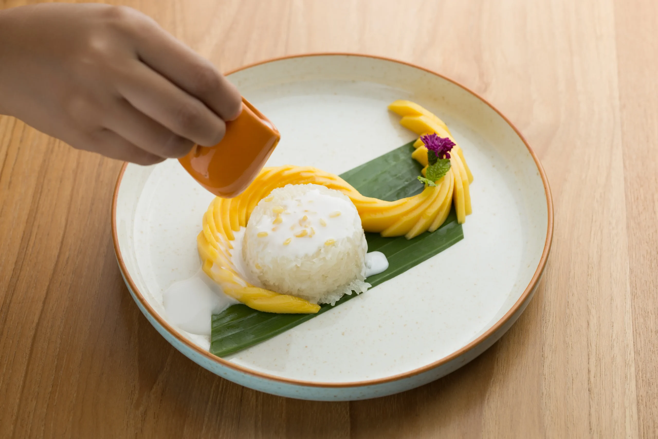 A traditional serving of sweet mango sticky rice topped with creamy coconut sauce, featuring ripe golden mango slices arranged beside glutinous rice and garnished with toasted sesame seeds on a rustic plate.