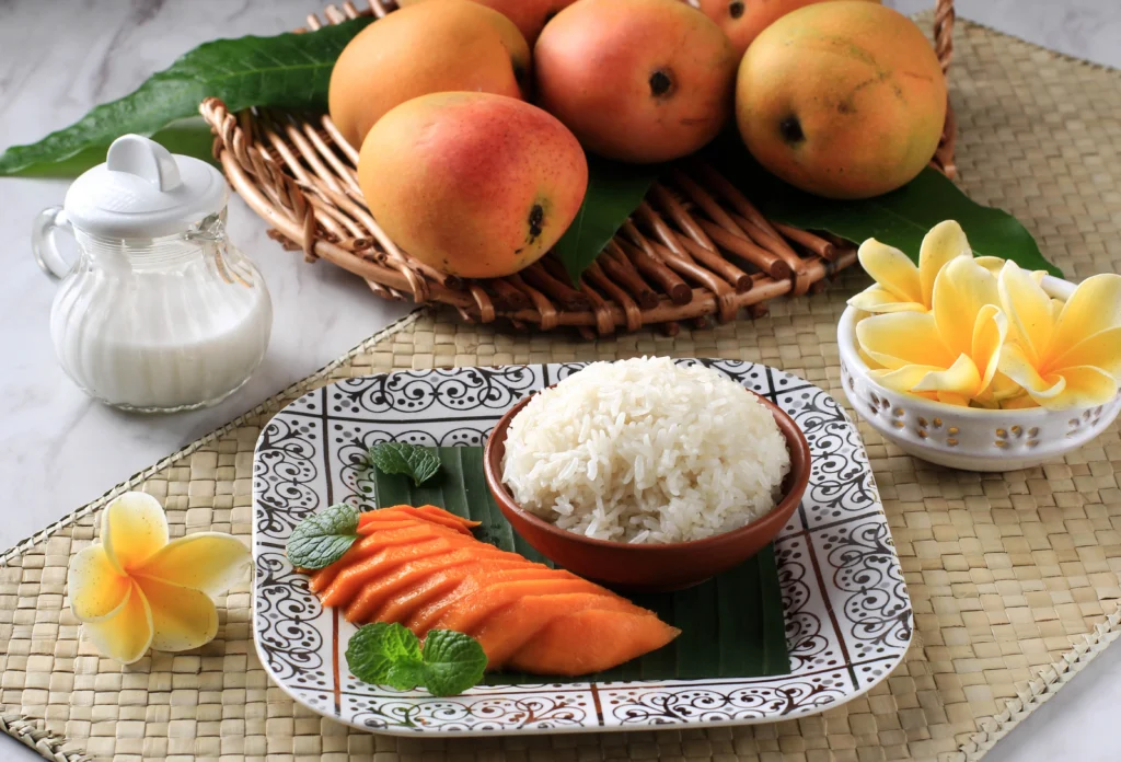A traditional serving of sweet mango sticky rice topped with creamy coconut sauce, featuring ripe golden mango slices arranged beside glutinous rice and garnished with toasted sesame seeds on a rustic plate.