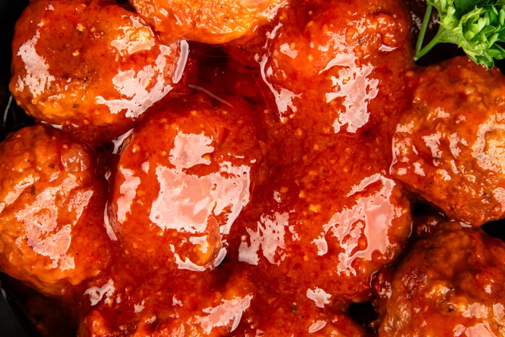 A bowl of sweet and sour meatballs coated in a glossy tangy sauce, garnished with fresh parsley and served alongside colorful bell peppers and pineapple chunks.