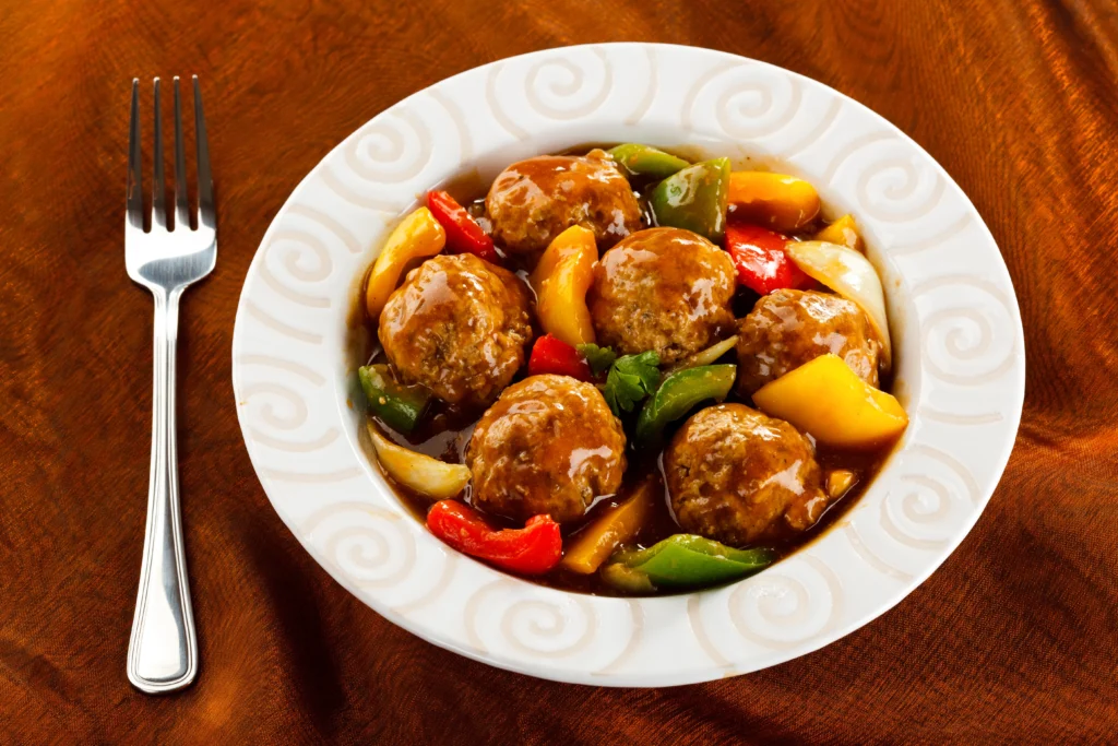 A bowl of sweet and sour meatballs coated in a glossy tangy sauce, garnished with fresh parsley and served alongside colorful bell peppers and pineapple chunks.