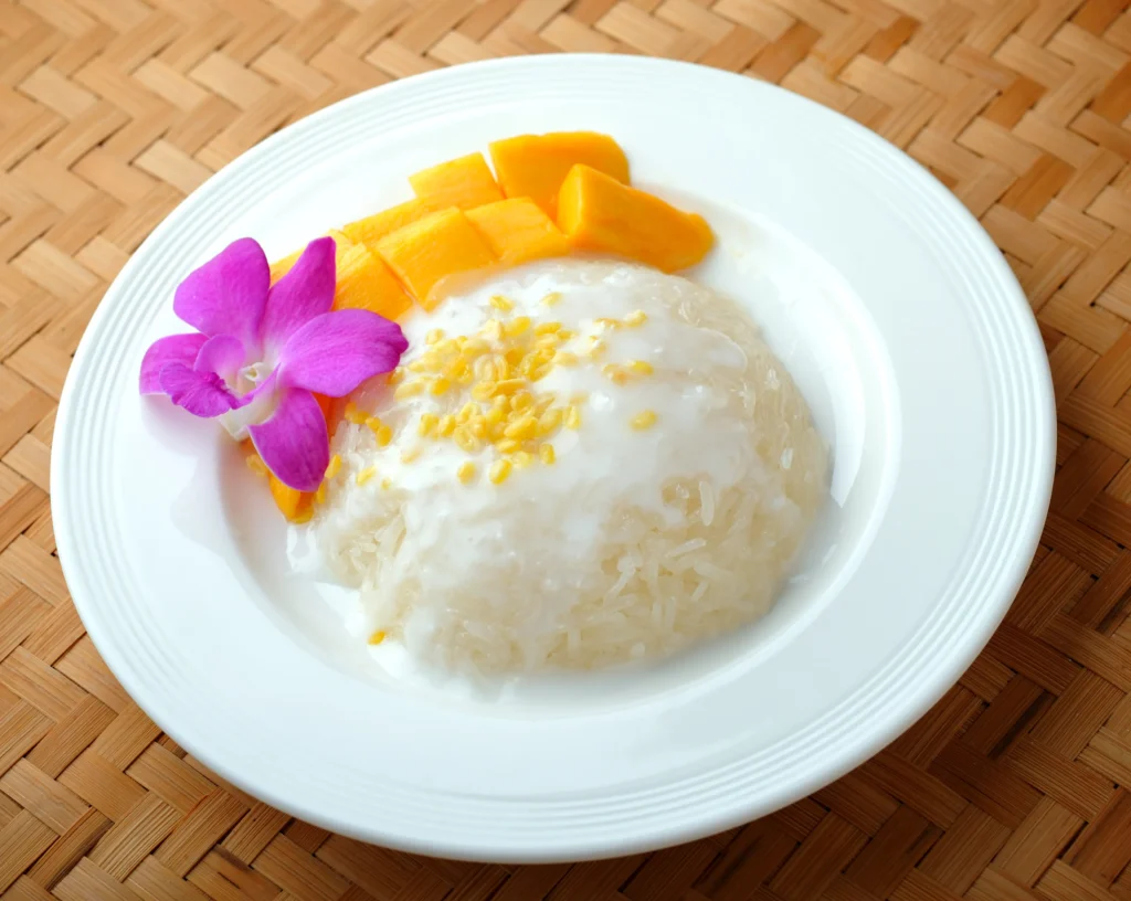A traditional serving of sweet mango sticky rice topped with creamy coconut sauce, featuring ripe golden mango slices arranged beside glutinous rice and garnished with toasted sesame seeds on a rustic plate.