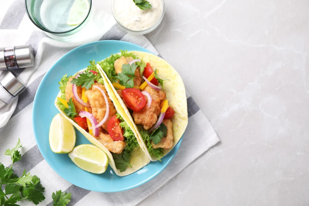 Fresh fish tacos served in warm corn tortillas with crispy white fish, shredded cabbage, avocado slices, pico de gallo, cilantro, lime wedges, and a creamy cilantro-lime sauce.