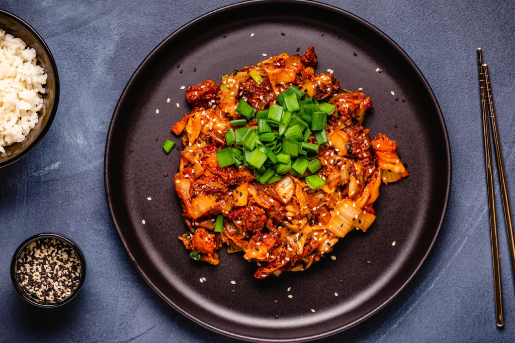 A colorful bowl of gochujang chicken stir-fry featuring tender chicken pieces coated in a spicy Korean gochujang sauce, stir-fried with bell peppers, broccoli, carrots, onions, and sesame seeds, served over steamed rice with sliced green onions and chopsticks.