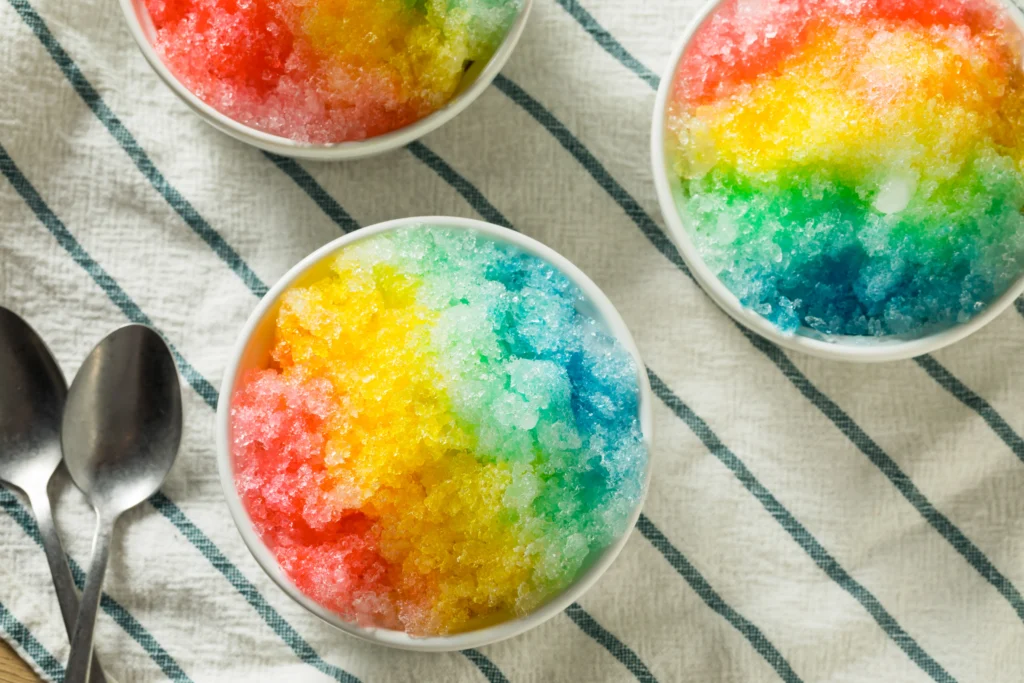 Homemade snow cone syrup in colorful bottles with shaved ice, fresh fruit, and summer toppings on a wooden table.