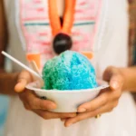 Homemade snow cone syrup in colorful bottles with shaved ice, fresh fruit, and summer toppings on a wooden table.