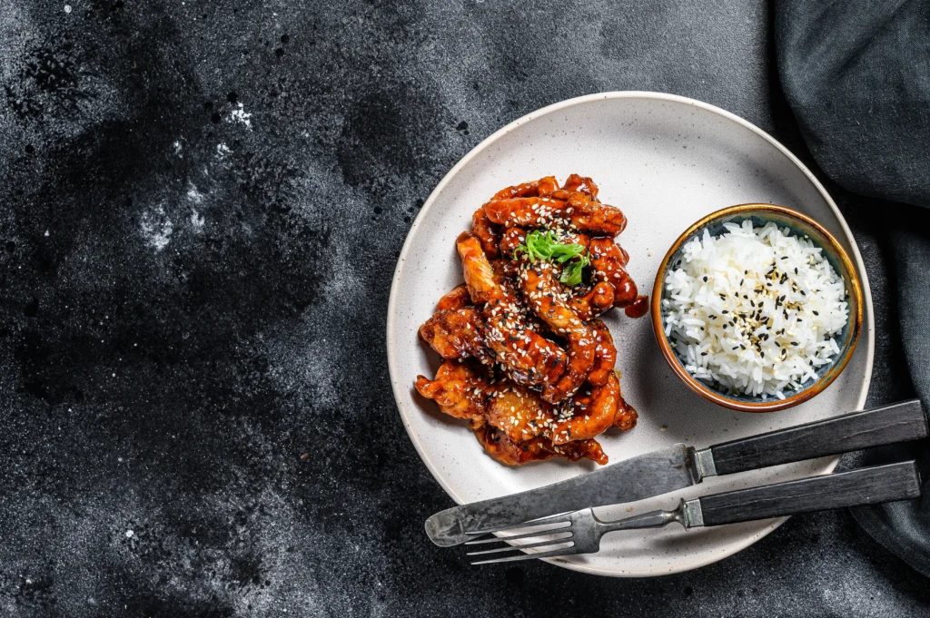 A colorful bowl of gochujang chicken stir-fry featuring tender chicken pieces coated in a spicy Korean gochujang sauce, stir-fried with bell peppers, broccoli, carrots, onions, and sesame seeds, served over steamed rice with sliced green onions and chopsticks.