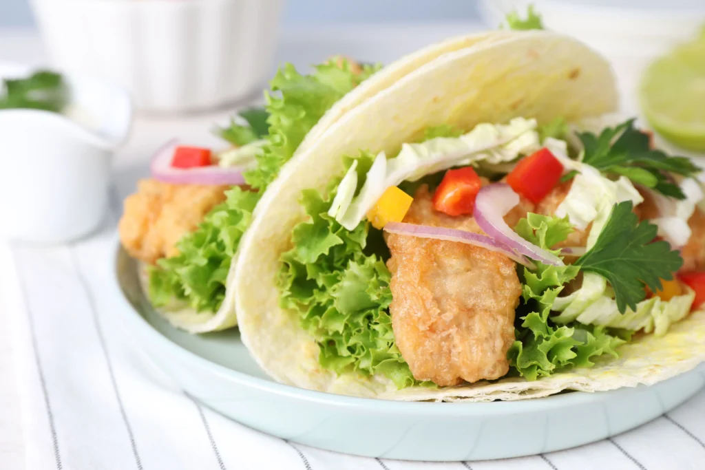 Fresh fish tacos served in warm corn tortillas with crispy white fish, shredded cabbage, avocado slices, pico de gallo, cilantro, lime wedges, and a creamy cilantro-lime sauce.