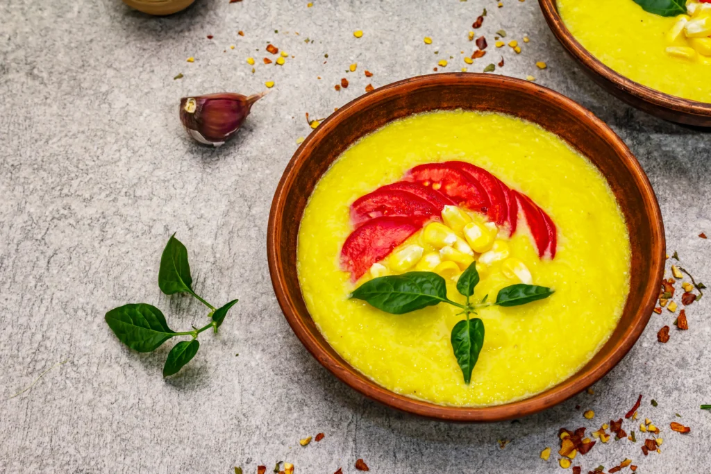 A bowl of creamy cold sweet corn gazpacho topped with fresh corn kernels, herbs, and a drizzle of olive oil, served chilled with crusty bread on a rustic table.